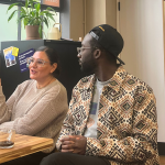 A woman in a beige sweater speaks animatedly while sitting at a wooden table beside a man in a patterned jacket and backwards baseball cap. Behind them, a black filing cabinet holds a plant and several posters, one of which reads, 