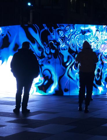 A crowd of people stands in a plaza at night, watching a large, curved LED screen. The screen is displaying a vibrant, abstract light show with swirling patterns in shades of electric blue and white. The figures in the foreground are silhouetted against the bright light of the screen, with the one in the center reaching out as if to touch it. In the background, there are other people, buildings, and a few small, glowing spotlights on the ground.