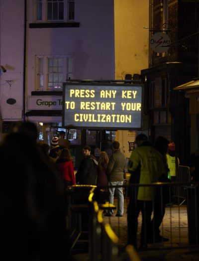 A large illuminated roadside sign on a trailer displays the words “PRESS ANY KEY TO RESTART CIVILZATION” in bright orange letters against a dark background at night.