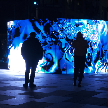 A crowd of people stands in a plaza at night, watching a large, curved LED screen. The screen is displaying a vibrant, abstract light show with swirling patterns in shades of electric blue and white. The figures in the foreground are silhouetted against the bright light of the screen, with the one in the center reaching out as if to touch it. In the background, there are other people, buildings, and a few small, glowing spotlights on the ground.