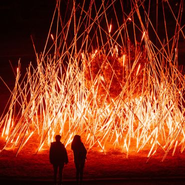 Two people look at a light installation at night - the artwork resembles a bonfire.