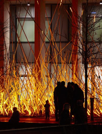 A dense cluster of long, thin poles glowing bright orange-red. The illuminated poles crisscross upwards at sharp angles, creating the effect of fiery sparks shooting from the ground. The artwork looks like a bonfire.